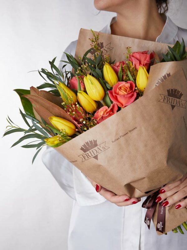 Bouquet Valencia, mazzo di fiori incartato con tulipani gialli e rose arancio. Ragazza con camicia bianca tiene in mano un bouquet di fiori con incarto Frida's