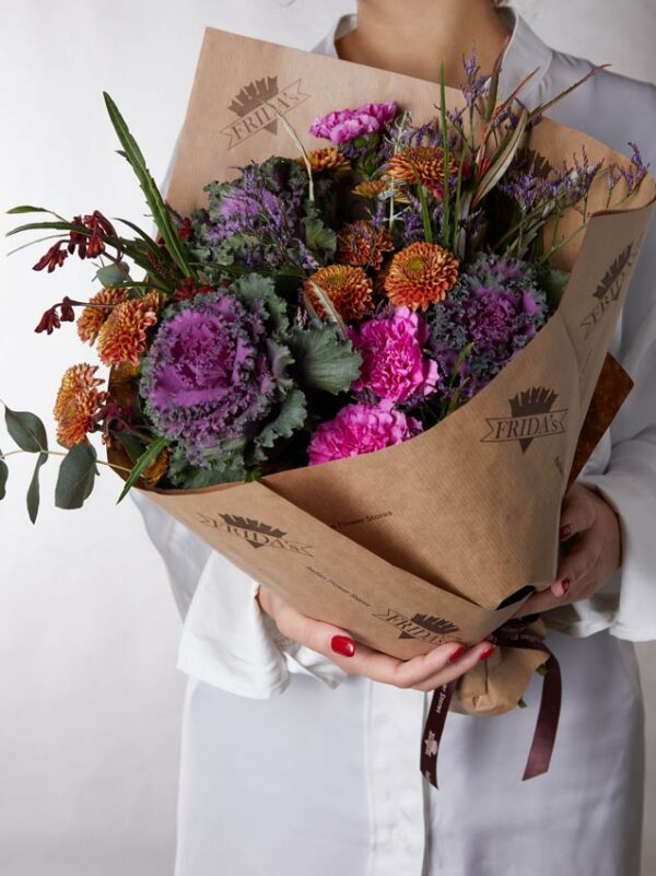 Bouquet Winter Rock, mazzo di fiori incartato con brassica, garofani fucsia e bottoncini. Ragazza con camicia bianca tiene in mano un bouquet di fiori con incarto Frida's