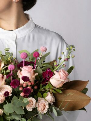 Bouquet Praga, mazzo di fiori incartato con tulipani fucsia, limonium e rose rosse. Ragazza con camicia bianca tiene in mano un bouquet di fiori con incarto Frida's