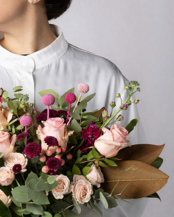 Bouquet Praga, mazzo di fiori incartato con tulipani fucsia, limonium e rose rosse. Ragazza con camicia bianca tiene in mano un bouquet di fiori con incarto Frida's